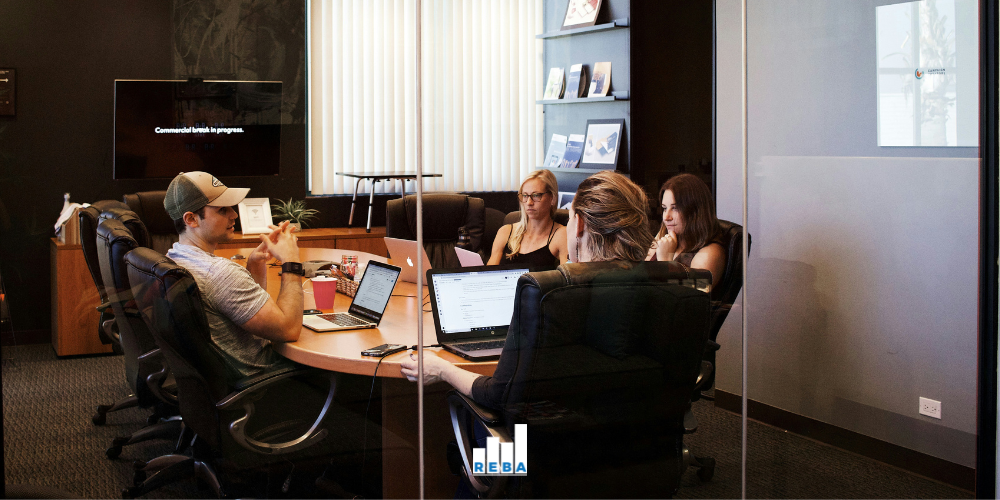 Four people sit around a conference table with laptops and papers in a modern office meeting room, viewed through glass walls. A TV screen and bookshelves are visible in the background.