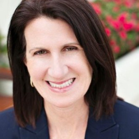 A woman with straight, dark brown hair and light skin smiles at the camera. She is wearing a navy blazer, giving her a polished and professional look that suggests a background in marketing. The blurred background features red flowers and greenery.
