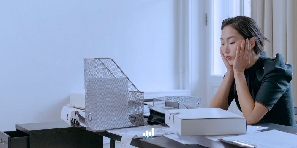 A woman sits at a cluttered desk, resting her face in her hands and looking tired or stressed, surrounded by papers, trays, and office supplies in a bright room.