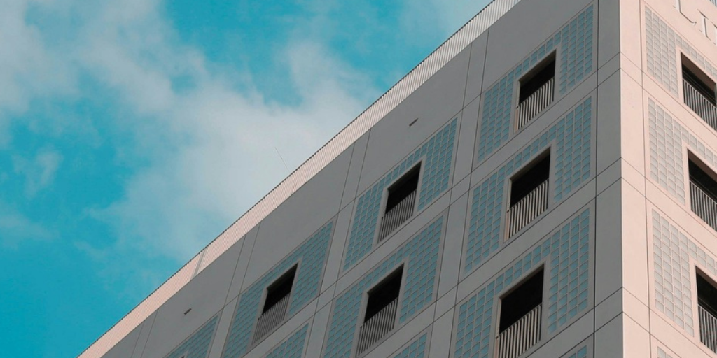 A beige building with square windows and railings is seen from below against a blue sky with clouds.
