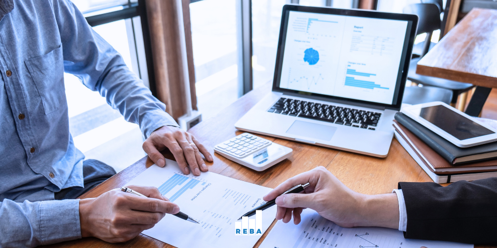 Two people discuss graphs and charts at a desk with documents, a calculator, a pen, and an open laptop displaying blue financial graphs.