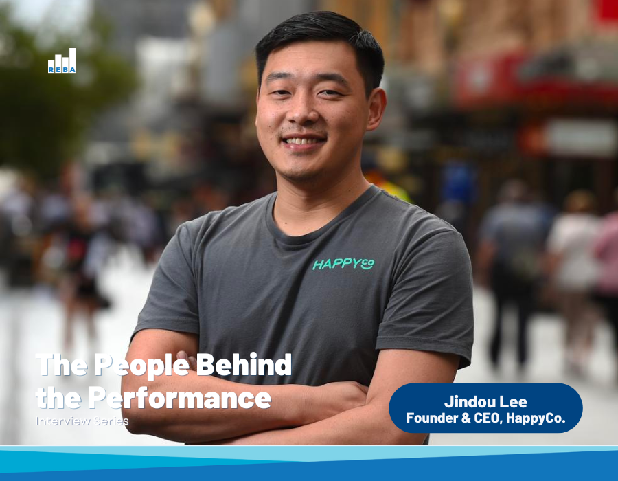 A man in a gray “HAPPYCO” shirt stands smiling with arms crossed on a busy city street. Text reads: “The People Behind the Performance: Jindou Lee, Founder & CEO, HappyCo.”.