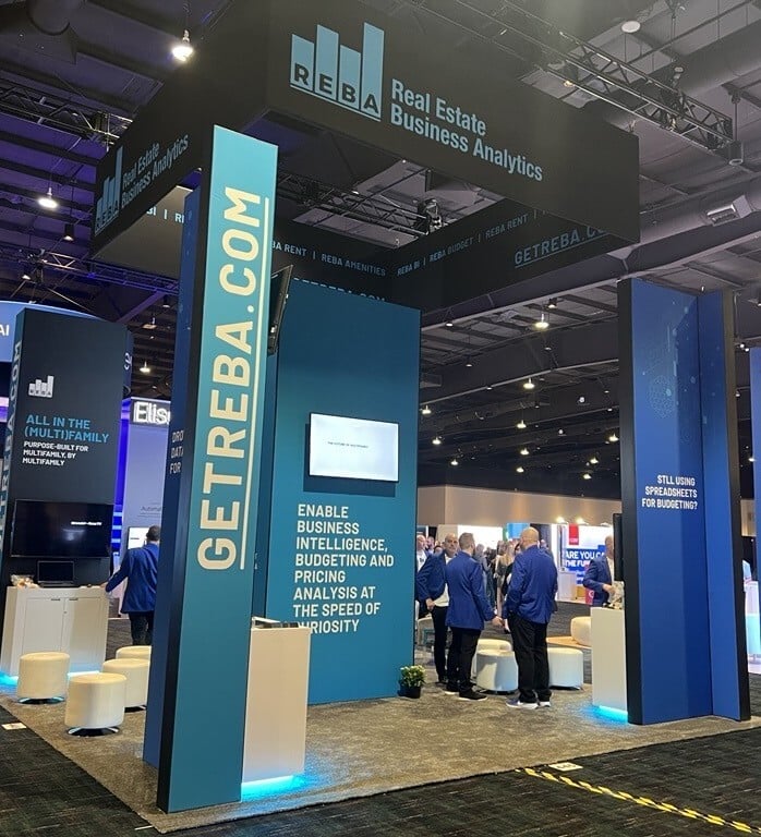 A trade show booth for Real Estate Business Analytics (REBA) with blue displays, seating, and people interacting. Signs promote business intelligence, budgeting, and pricing analysis tools.