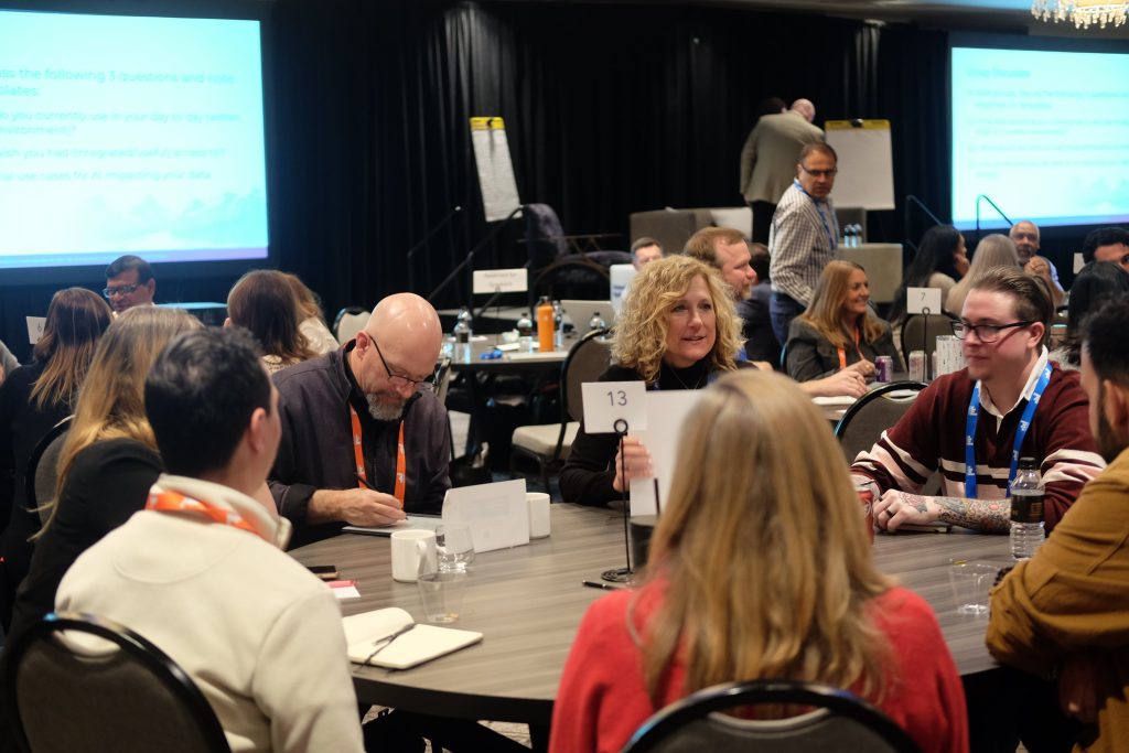 People seated around round tables at a conference, engaging in discussion. A woman holds up a numbered sign. The room has presentation screens, people taking notes, and a collaborative atmosphere.