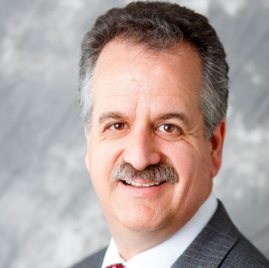 A man with gray hair and a mustache, wearing a gray suit and a red tie, smiles in front of a gray marbled background.
