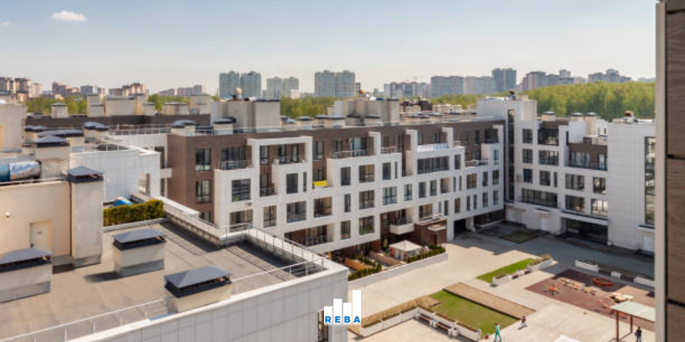 Modern apartment complex with multiple stories, rooftop structures, and a central courtyard. Other tall buildings and trees are visible in the background under a clear sky.