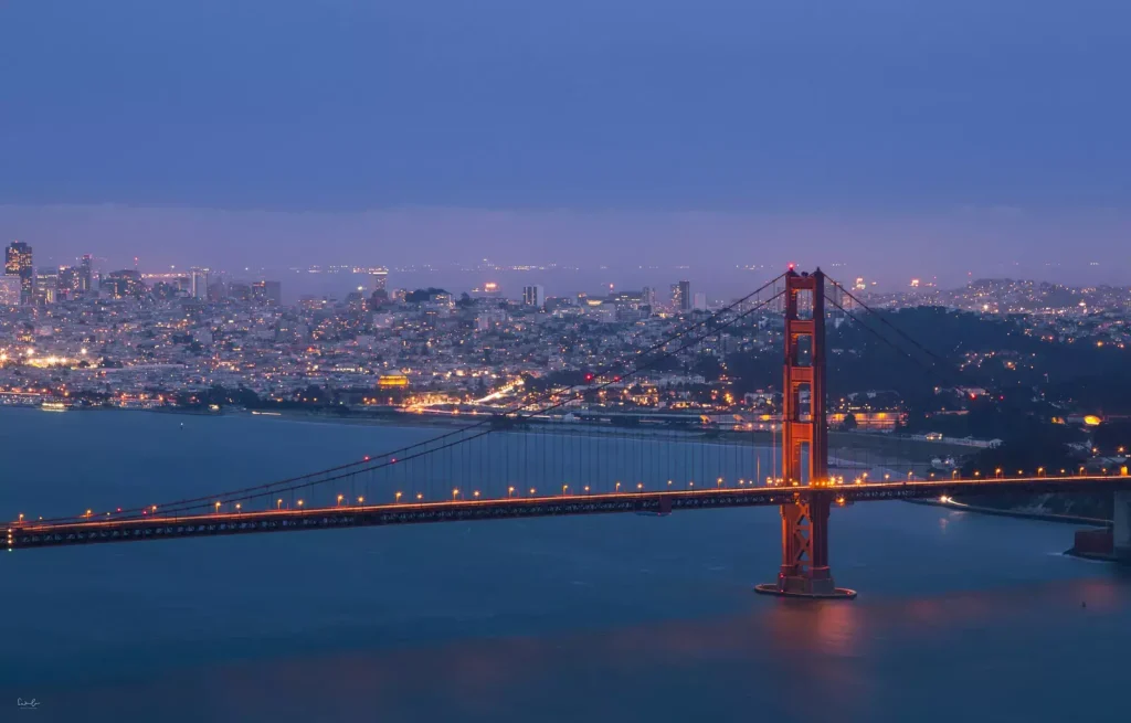 Golden Gate Bridge illuminated at dusk, with city lights of San Francisco in the background and the bay’s water in the foreground under a cloudy sky.