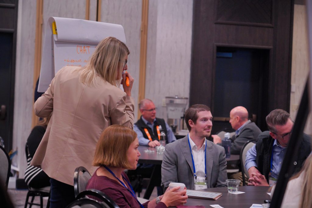 A woman writes on a large notepad while others sit and talk around tables in a conference room setting.