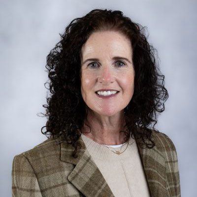 Woman with curly dark hair wearing a brown plaid blazer and light sweater, smiling at the camera against a plain light gray background.