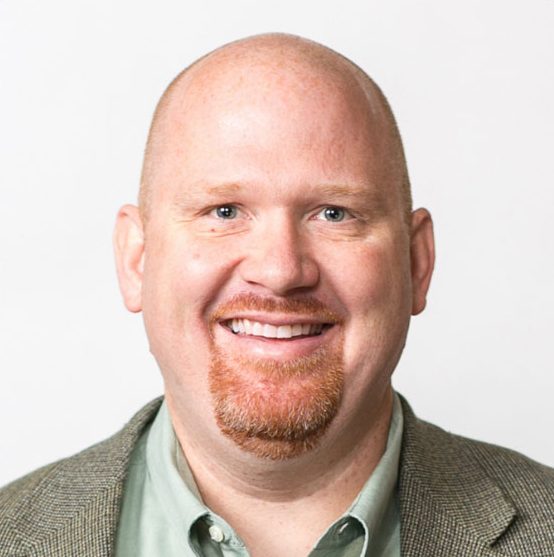 A smiling bald man with a reddish goatee, wearing a gray blazer over a green collared shirt, in front of a plain white background.