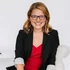 A person with shoulder-length light brown hair, wearing glasses, a black blazer, and a red top, sits on a white couch, smiling at the camera.