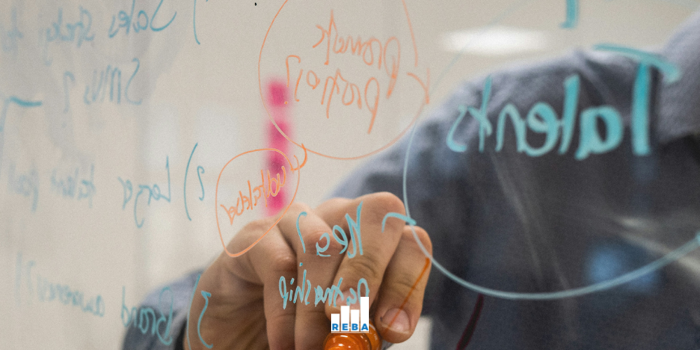 A person writes colorful notes and diagrams on a transparent glass board using an orange marker, with words and arrows visible through the board.