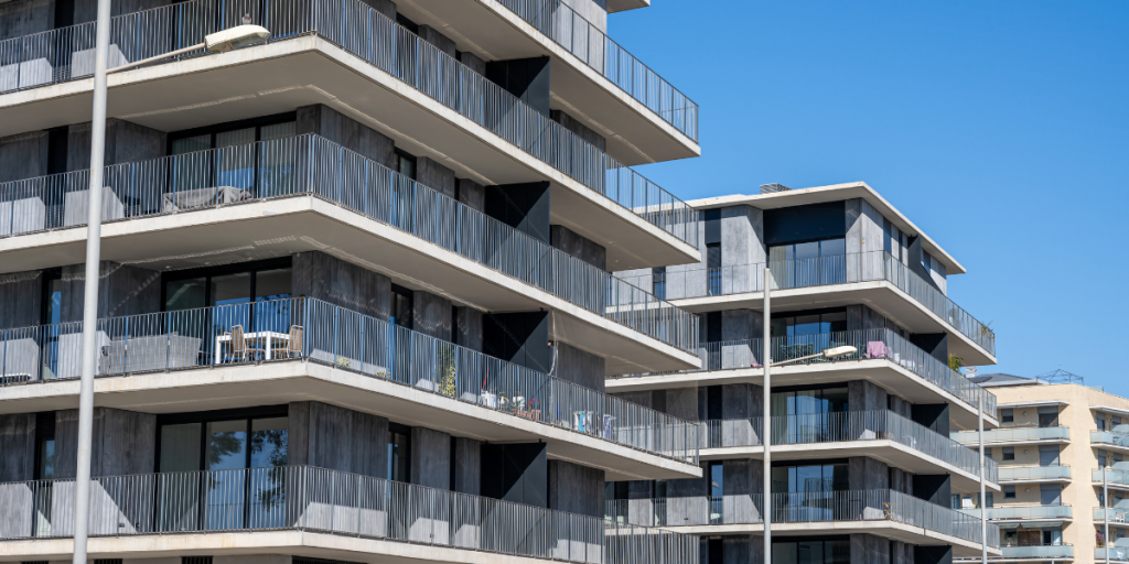Modern apartment buildings with large glass windows and multiple balconies on each floor, set against a clear blue sky on a sunny day.