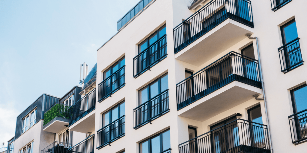 Modern apartment building with multiple floors, large windows, and black metal railings on each balcony. The facade is light-colored, and the sky above is clear and blue.