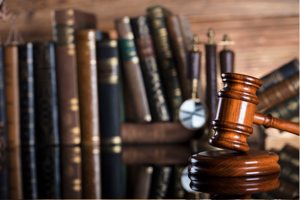 A wooden judge’s gavel rests on a sound block, with blurred law books and a balance scale in the background, symbolizing justice and legal proceedings.