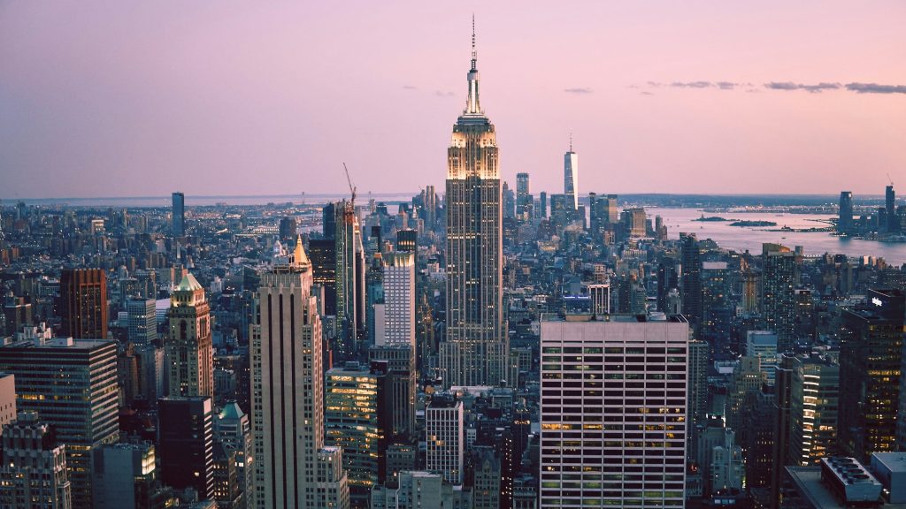 New York City skyline at sunset with the Empire State Building lit up in the center, surrounded by skyscrapers and a pastel sky in the background.