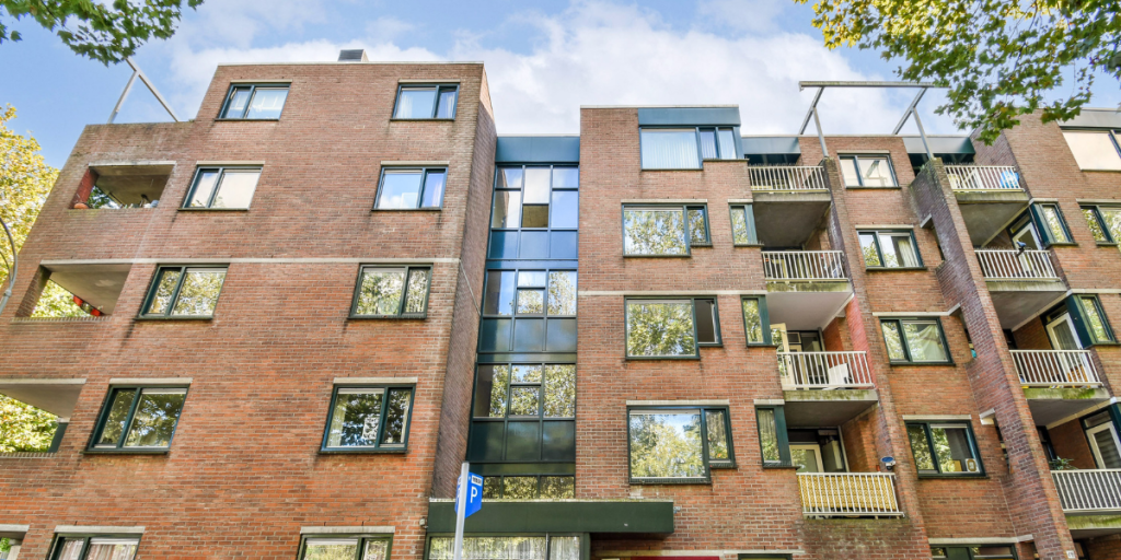 A brick apartment building with multiple balconies and large windows, viewed from below. Trees and blue sky are reflected in the windows, and a blue sign is visible near the entrance.