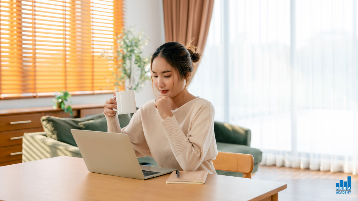 A woman sits at a table with a laptop and notebook, holding a white mug. She appears focused, in a bright room with large windows, a green sofa, and wooden blinds.