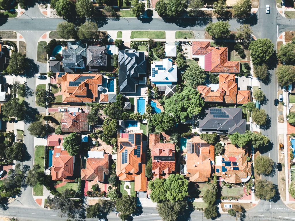 Aerial view of a suburban neighborhood with houses, trees, and swimming pools. The rooftops vary in color, and the streets surround the houses on all sides.