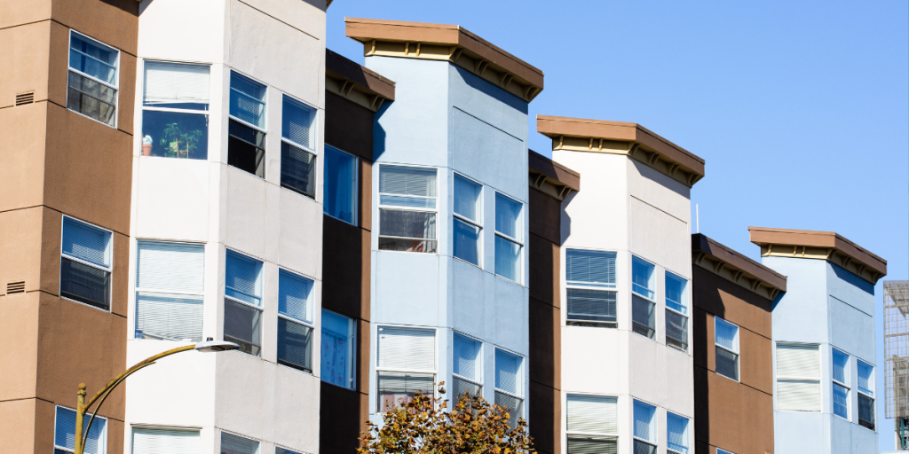 Modern apartment building with alternating brown and blue sections, each featuring large windows with white blinds. The clear blue sky is in the background, and a tree and streetlamp are visible in the foreground.