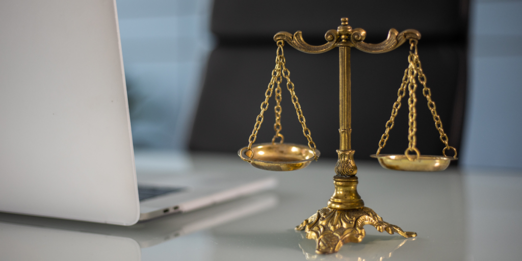 A brass balance scale sits on a glass desk next to a silver laptop, suggesting a legal or justice theme in an office setting.