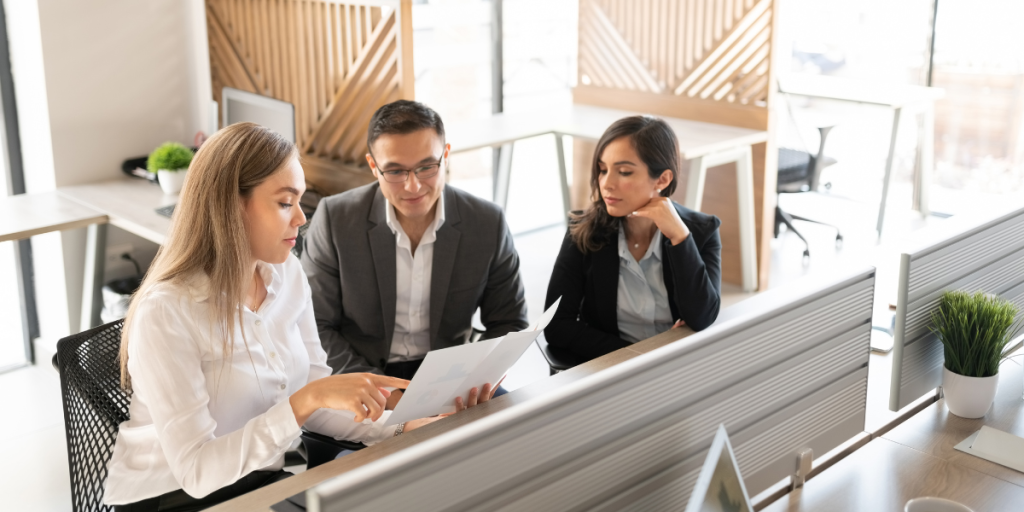 Three people in business attire sit at a desk in a modern office, discussing documents. One woman points to a paper while the others listen attentively. Computer monitors and plants are visible in the workspace.