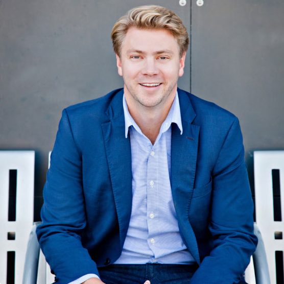 A man with blond hair in a blue suit and light blue shirt sits on a white bench, smiling at the camera, with a dark gray background behind him.