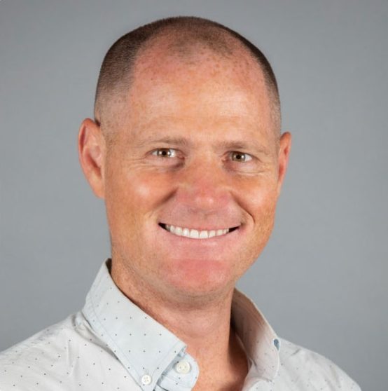A smiling man with short, closely-cropped hair wears a light-colored, button-up shirt with small dots, posing in front of a plain gray background.