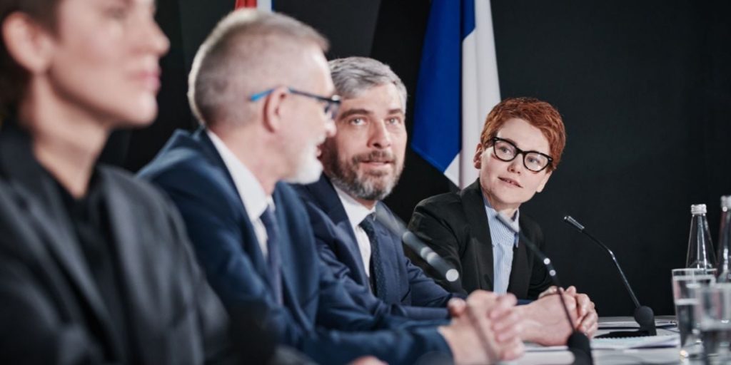 Four people in business attire sit at a conference table with microphones, engaged in discussion. Flags and water bottles are visible in the background.