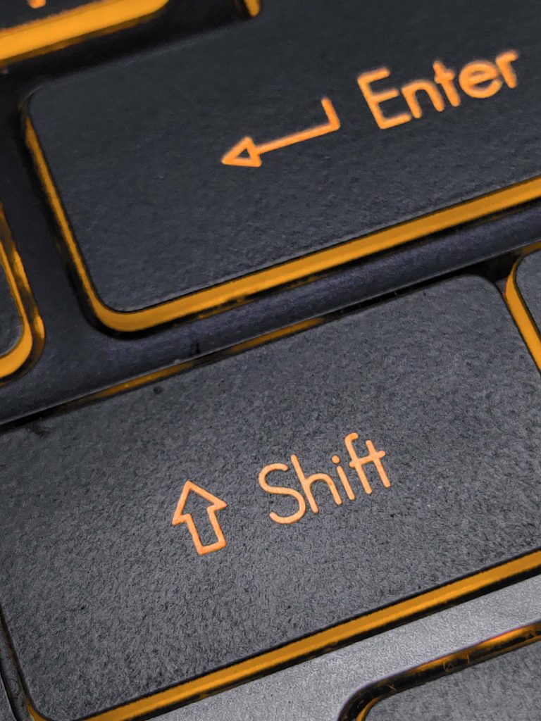 Close-up of a black keyboard with orange lettering, focusing on the Shift key in the center and part of the Enter key above it. The keys have a bold arrow and text, with orange outlines between the keys.