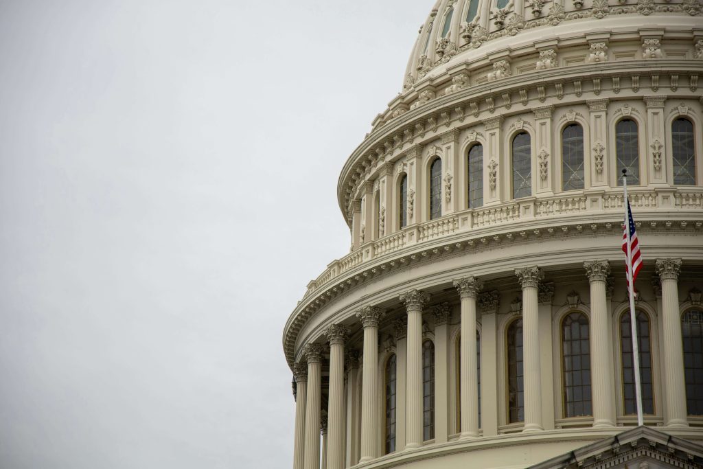 Close-up of the U.S. Capitol building’s dome, showing columns, windows, and part of an American flag against a cloudy sky.