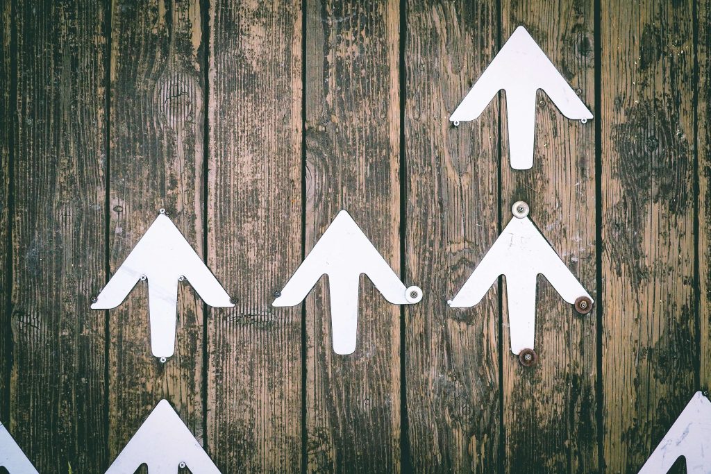 Five white arrows pointing upward are arranged on a weathered wooden surface, suggesting direction or movement. The arrows vary slightly in placement and orientation.