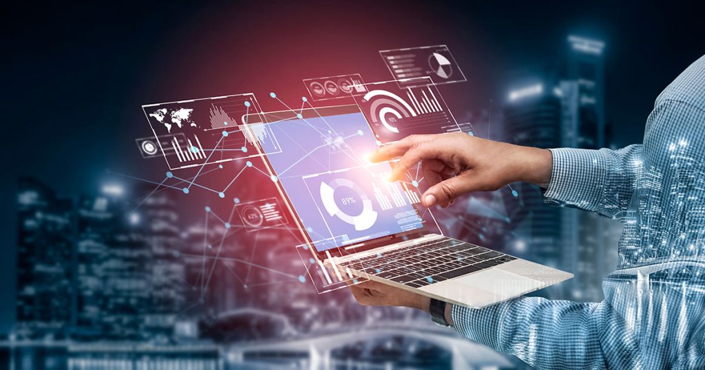 An analyst is using a laptop with virtual data graphics, charts, and world maps appearing above the screen, set against a blurred cityscape background at night.