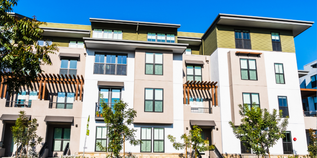 A modern apartment building with large windows, beige and green exterior walls, and wooden accents, set against a bright blue sky with trees in front.